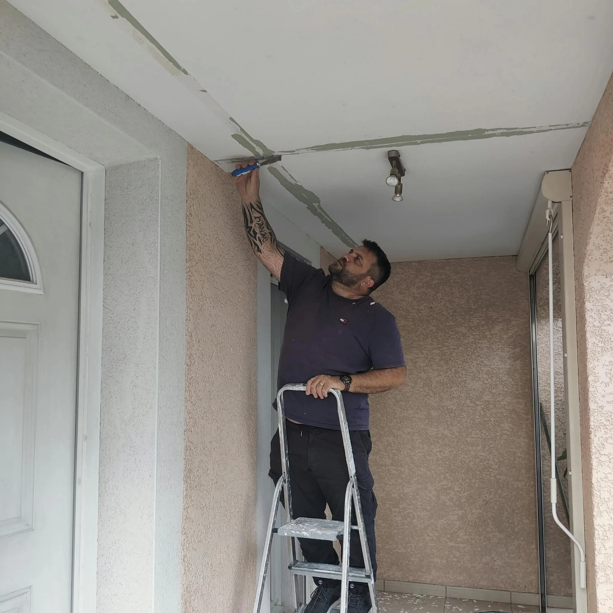 Artisan sur un escabeau grattant la peinture écaillée le long des joints d’un plafond de porche